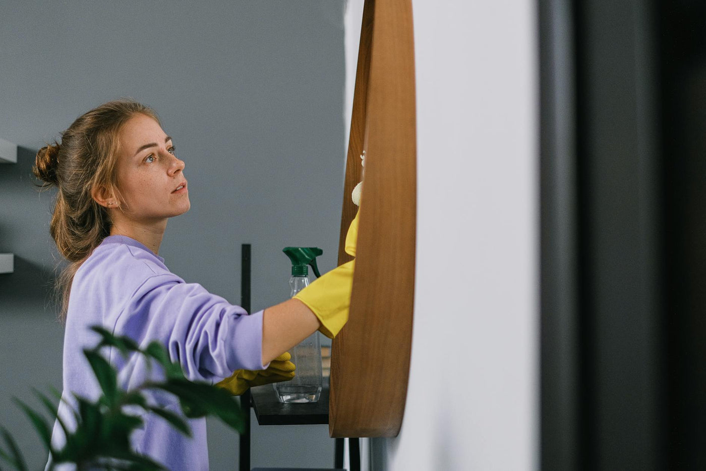 Side view of thoughtful young woman with brown hair in latex gloves cleaning round mirror in light room against gray background in daytime
